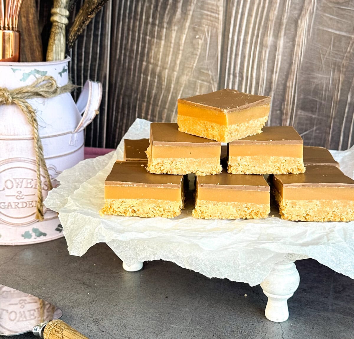 Caramel No Bake Slice on a white stand with a dark background and kitchen accessories 
