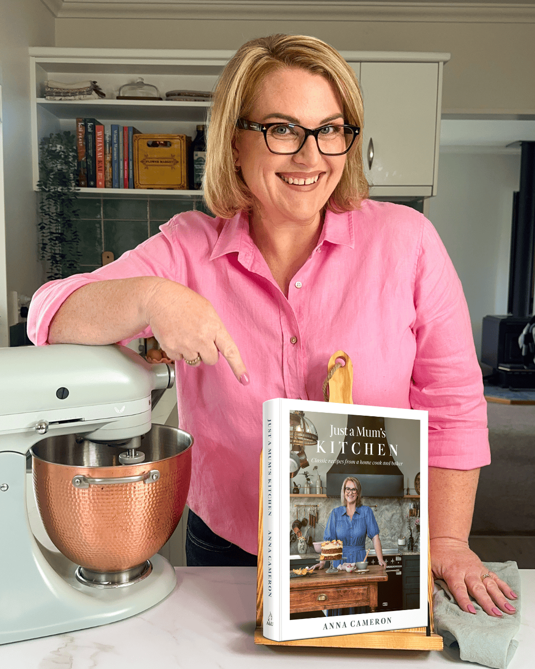 Anna Cameron with a mockup of her first cookbook on a stand for Just A Mum's Kitchen, standing in her own kitchen. 