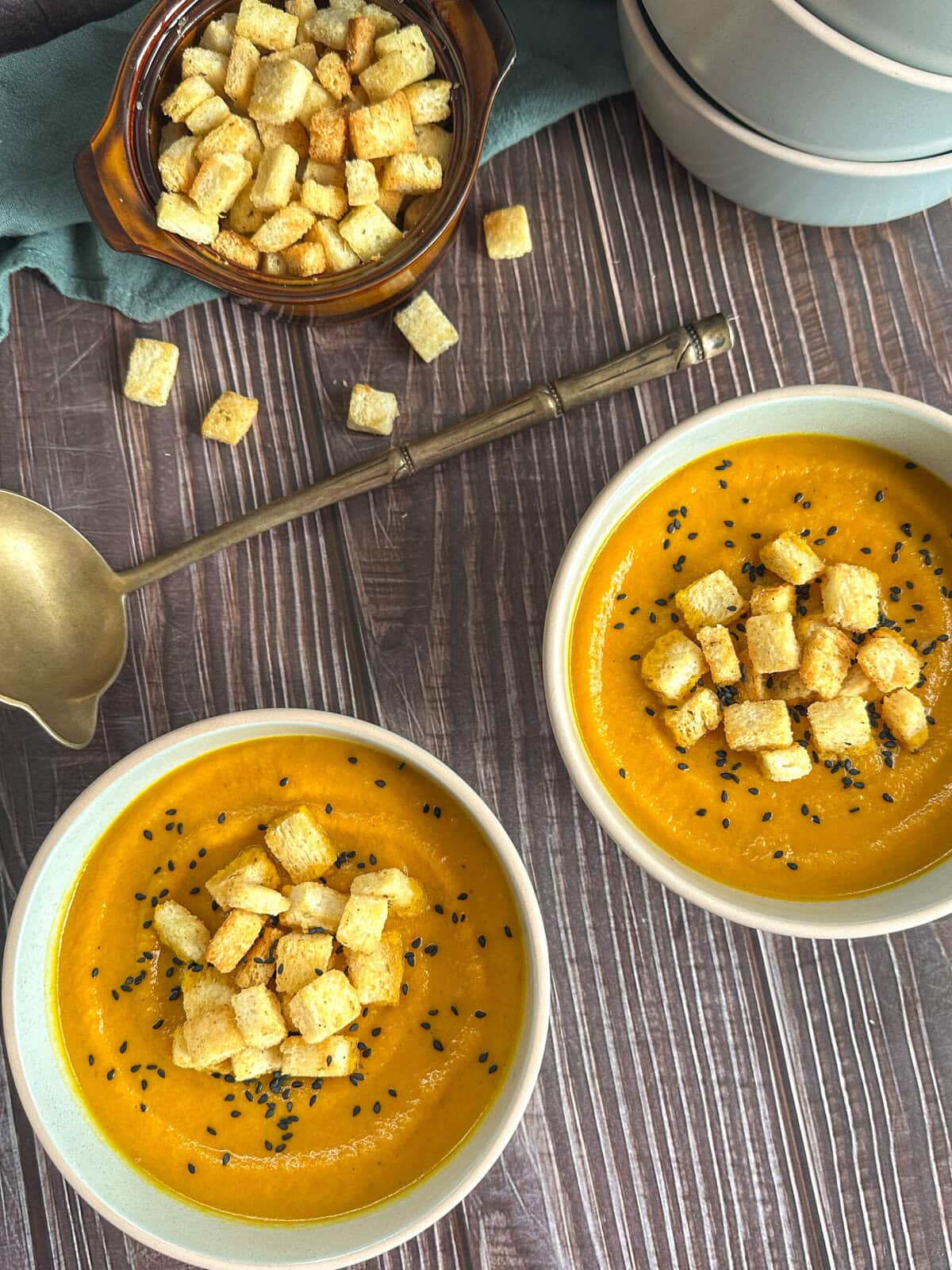 overhead photograph of two bowls of roasted carrot soup, croutons, a soup ladle and black sesame seeds