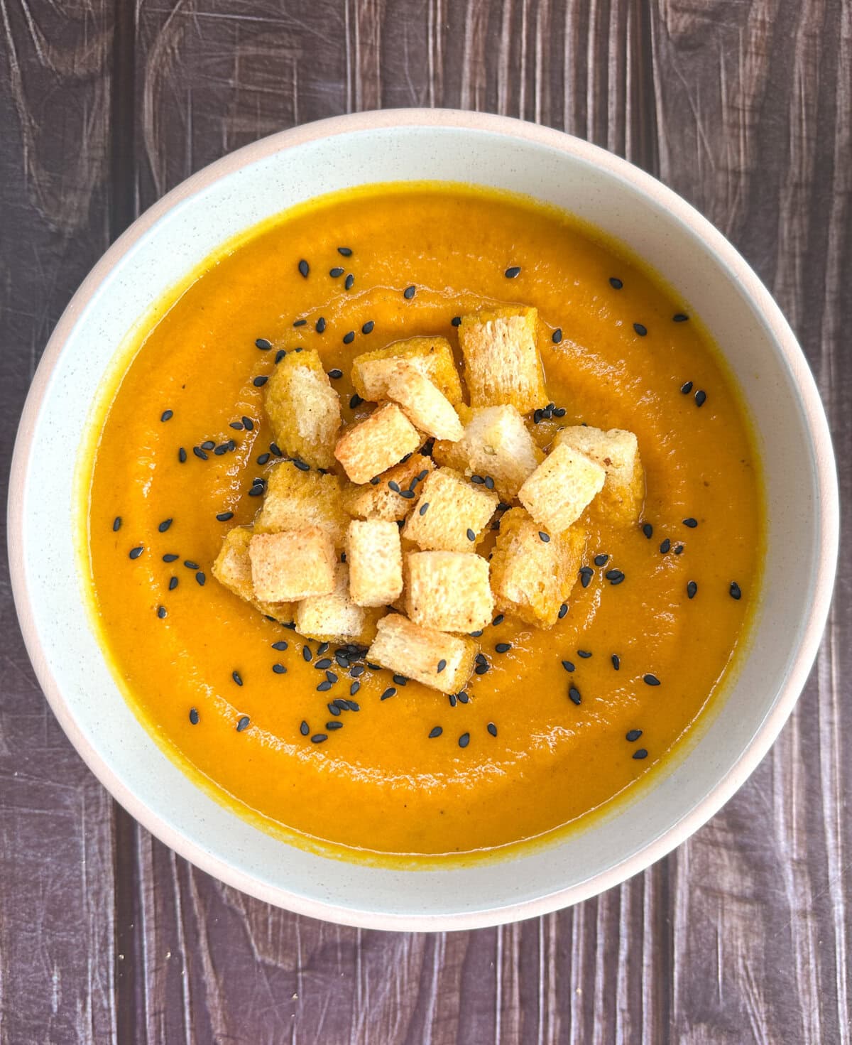 A pale green bowl with roasted carrot and cumin soup with croutons and black sesame seeds