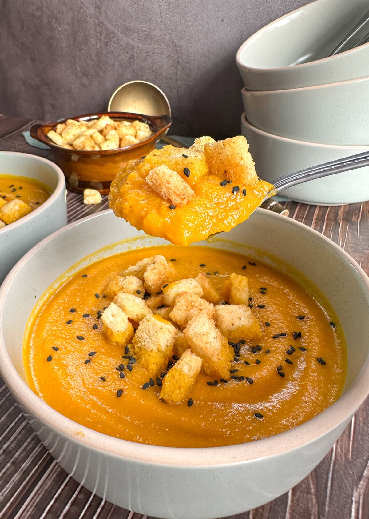 A soup spoon holding up a spoonful carrot soup with croutons with bowls in the background