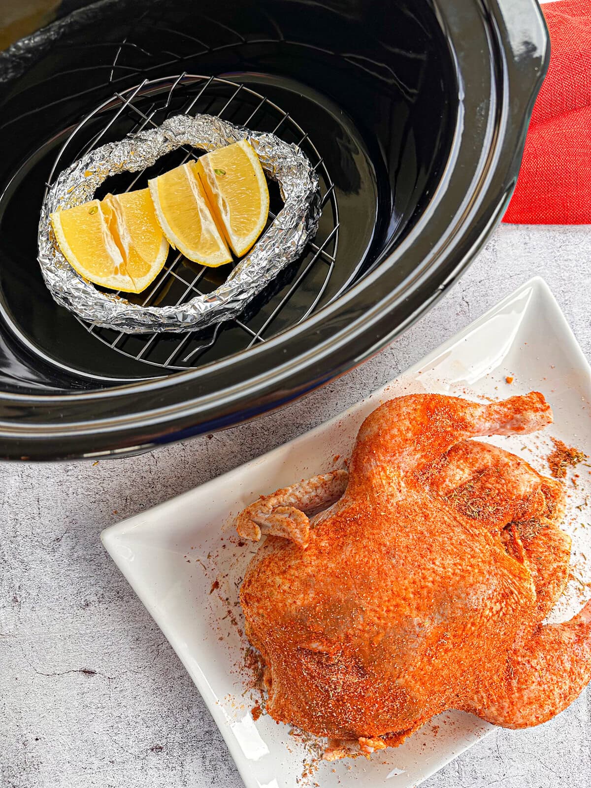 Raw chicken coated in a spice rub on a white plate with a slow cooker with foil, wire rack and lemons
