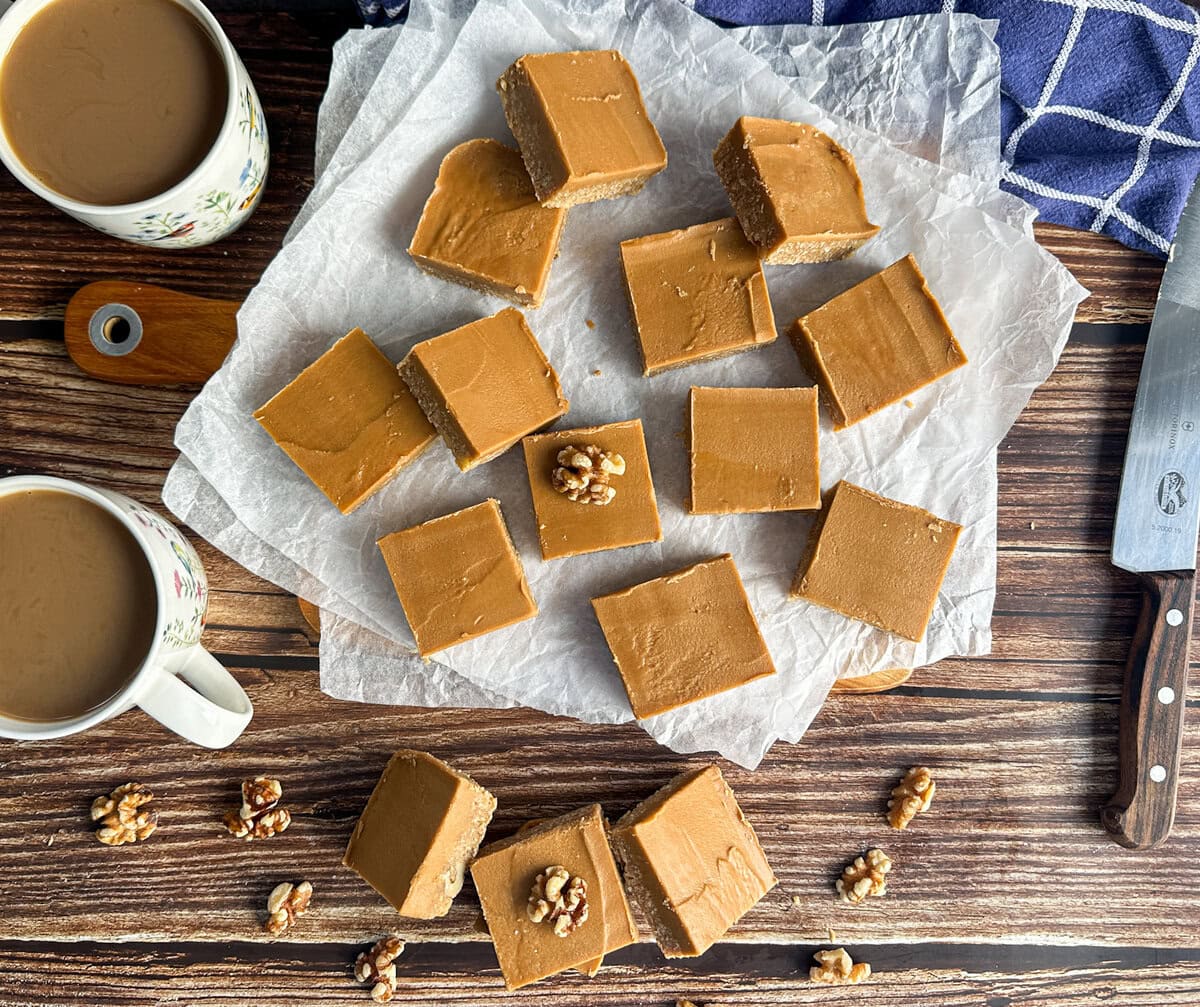 Pieces of No Bake Coffee and Walnut Fudge Slice on a wooden table with cups of coffee and a wooden knife in the background. 
