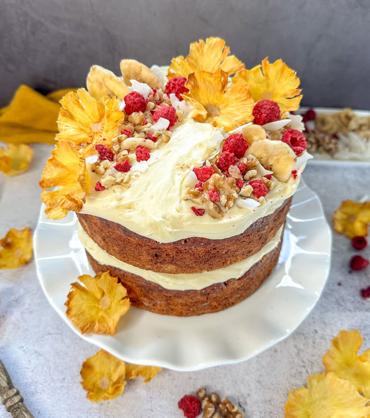 Beautiful Hummingbird Cake, two layers with cream cheese icing and dried pineapple flowers, freeze dried raspberries, coconut flakes, walnuts and dried banana as decoration on a white cake stand . 