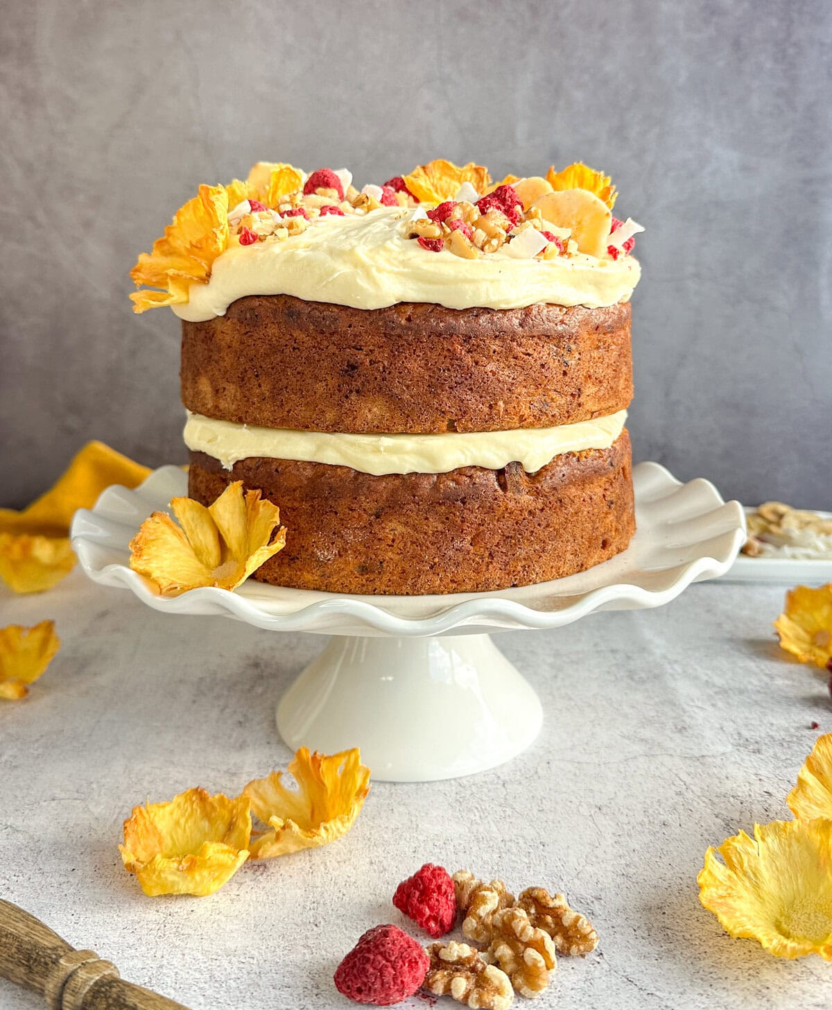 Hummingbird Cake, two layers with cream cheese icing and dried pineapple flowers, freeze dried raspberries, coconut flakes, walnuts and dried banana as decoration on a white cake stand . 
