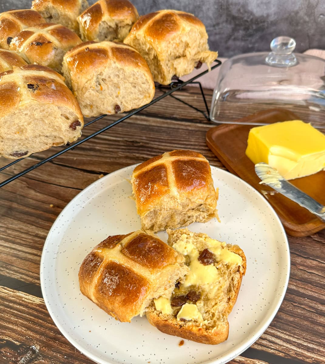 Hot Cross Buns halved with butter, served on a white plate with a tray of more buns in the background.
